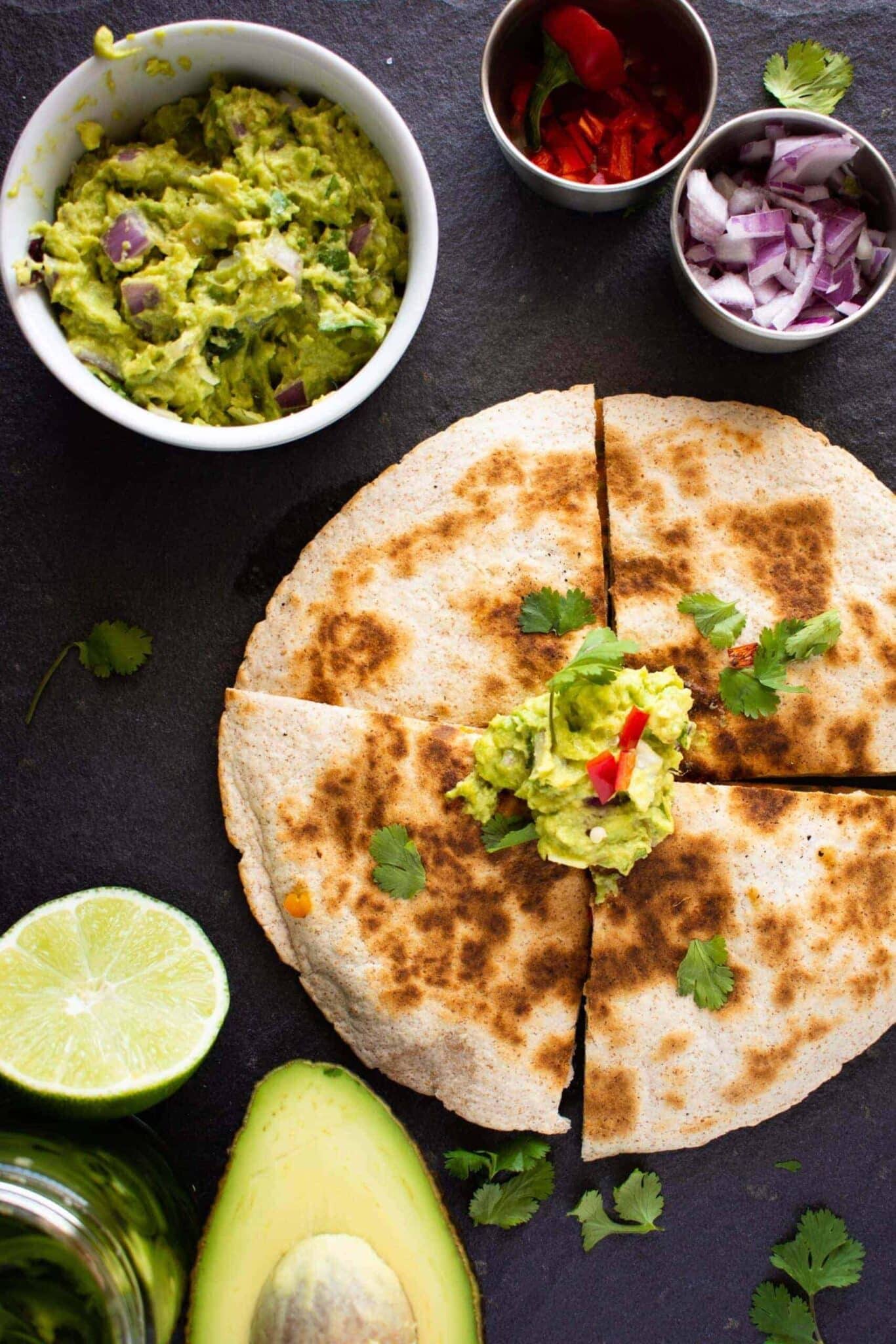 Vegan Butternut Squash Quesadillas with cilantro and guacamole, plus red onion, an avocado and lime on the table.