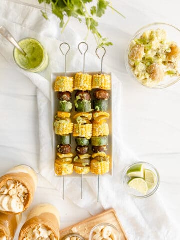 Grilled Veggie Kebabs - Corn, mushrooms, zucchini, and bell peppers on a metal skewer drizzled with chimichurri sauce, and accompanied by peanut butter banana pudding and potato salad.