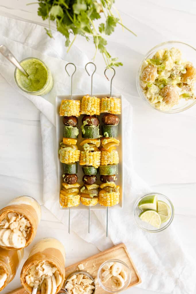 Grilled Veggie Kebabs - Corn, mushrooms, zucchini, and bell peppers on a metal skewer drizzled with chimichurri sauce, and accompanied by peanut butter banana pudding and potato salad. 