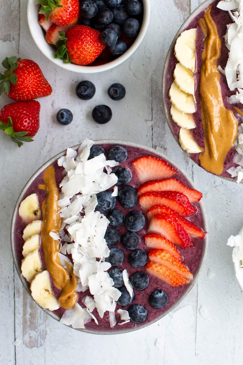 two thick berry smoothie bowls topped with bananas, peanut butter, coconut flakes, blueberries and strawberries