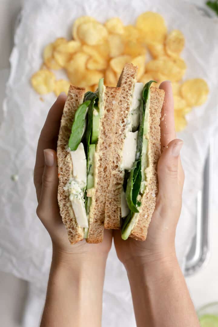 holding a vegan green goddess sandwich with potato chips in the background