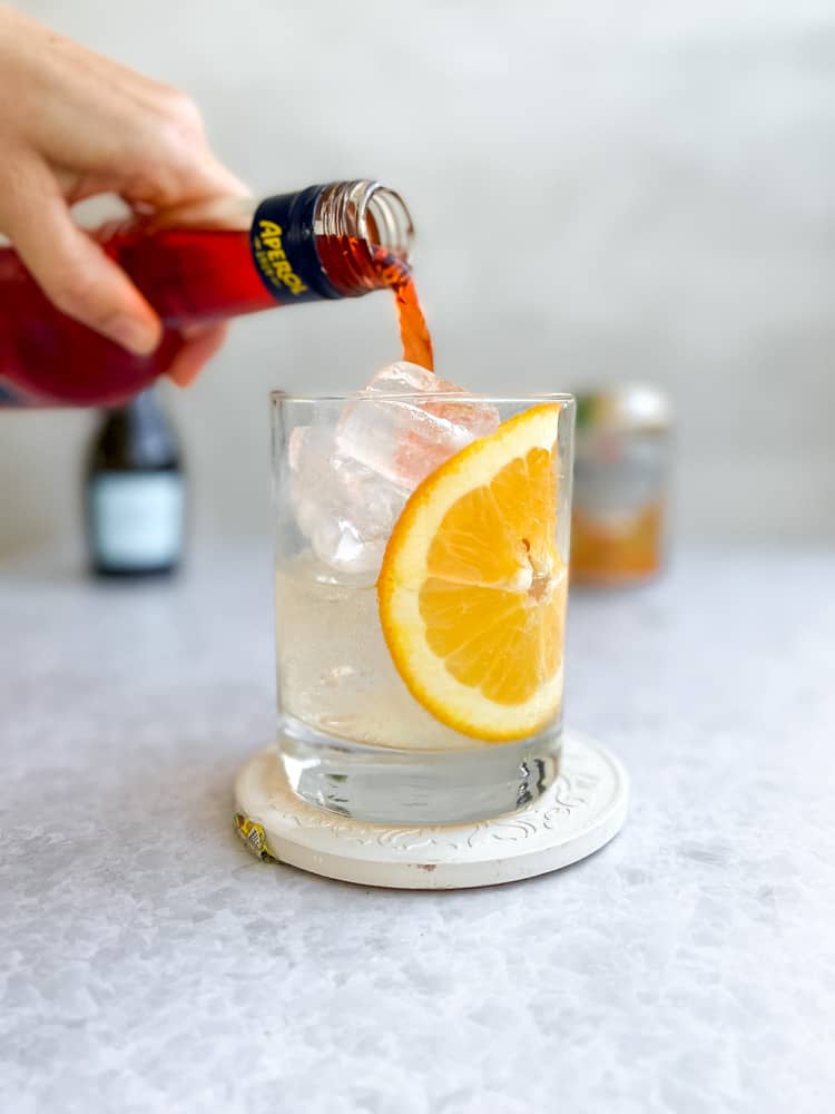 Pouring aperol into a glass with ice, an orange slice, and prosecco
