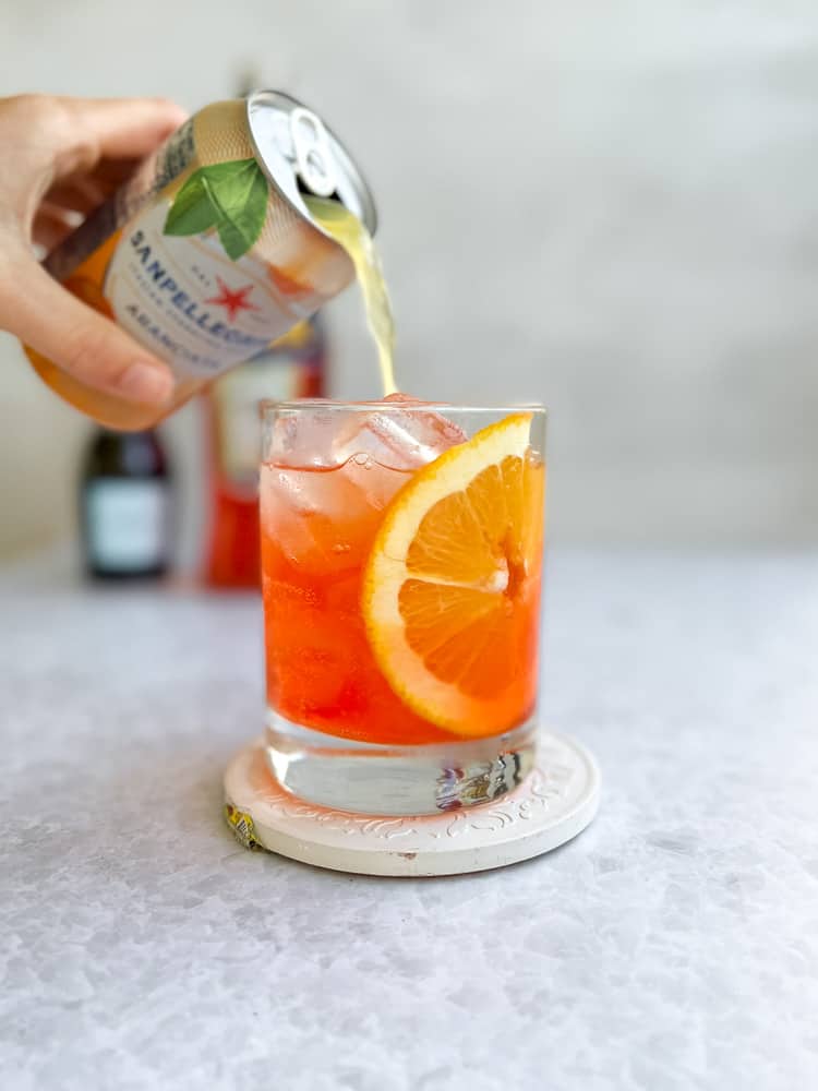 Pouring soda into a glass with ice, an orange slice, prosecco and aperol