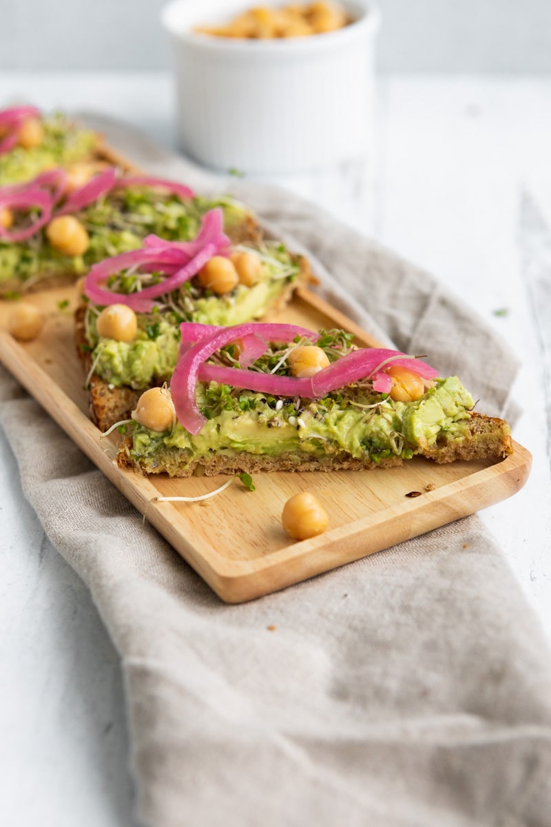 avocado toast with chickpeas and red pickled onion on a wooden platter next to a ramekin with cooked chickpeas.