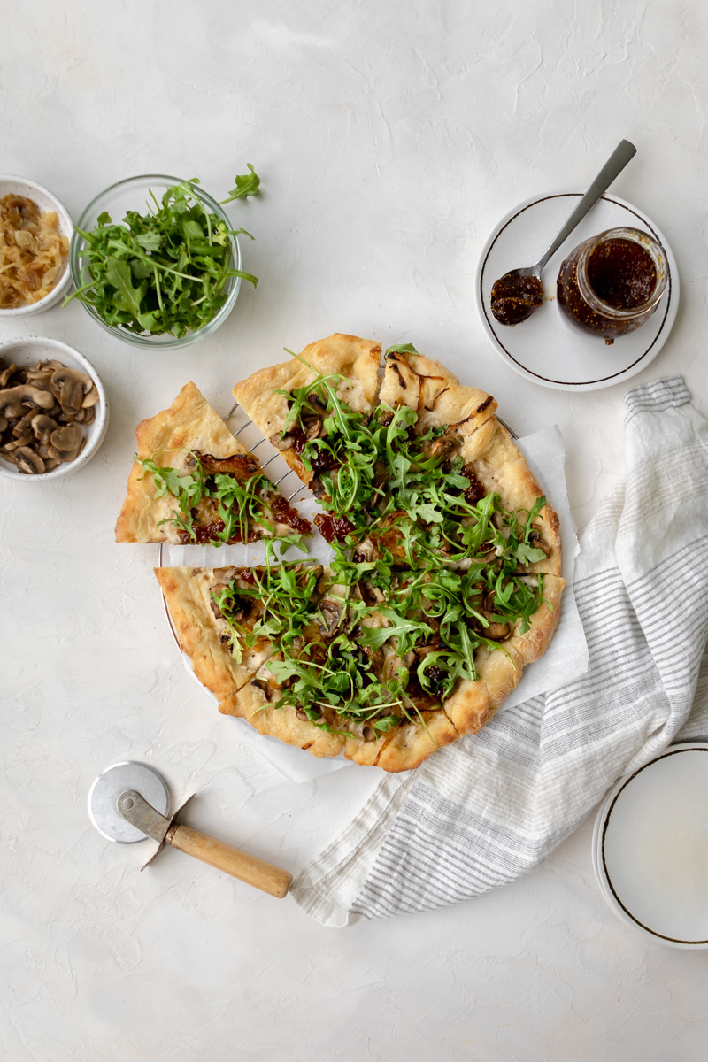 Slice of dairy free pizza with arugula, fig jam, white sauce, caramelized onions and mushrooms 
