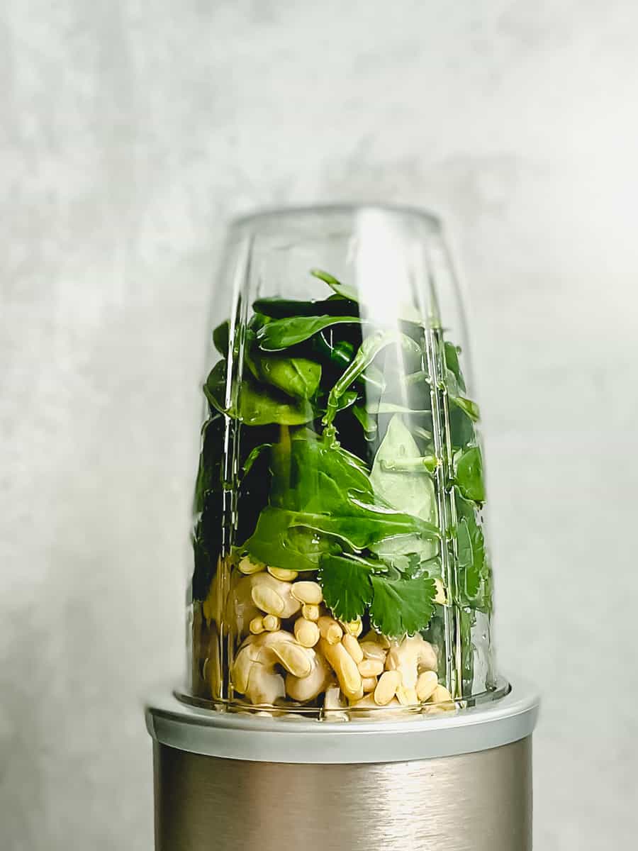 pesto ingredients in a personal blender, with cashews on the bottom, followed by leafy greens and herbs on top. 