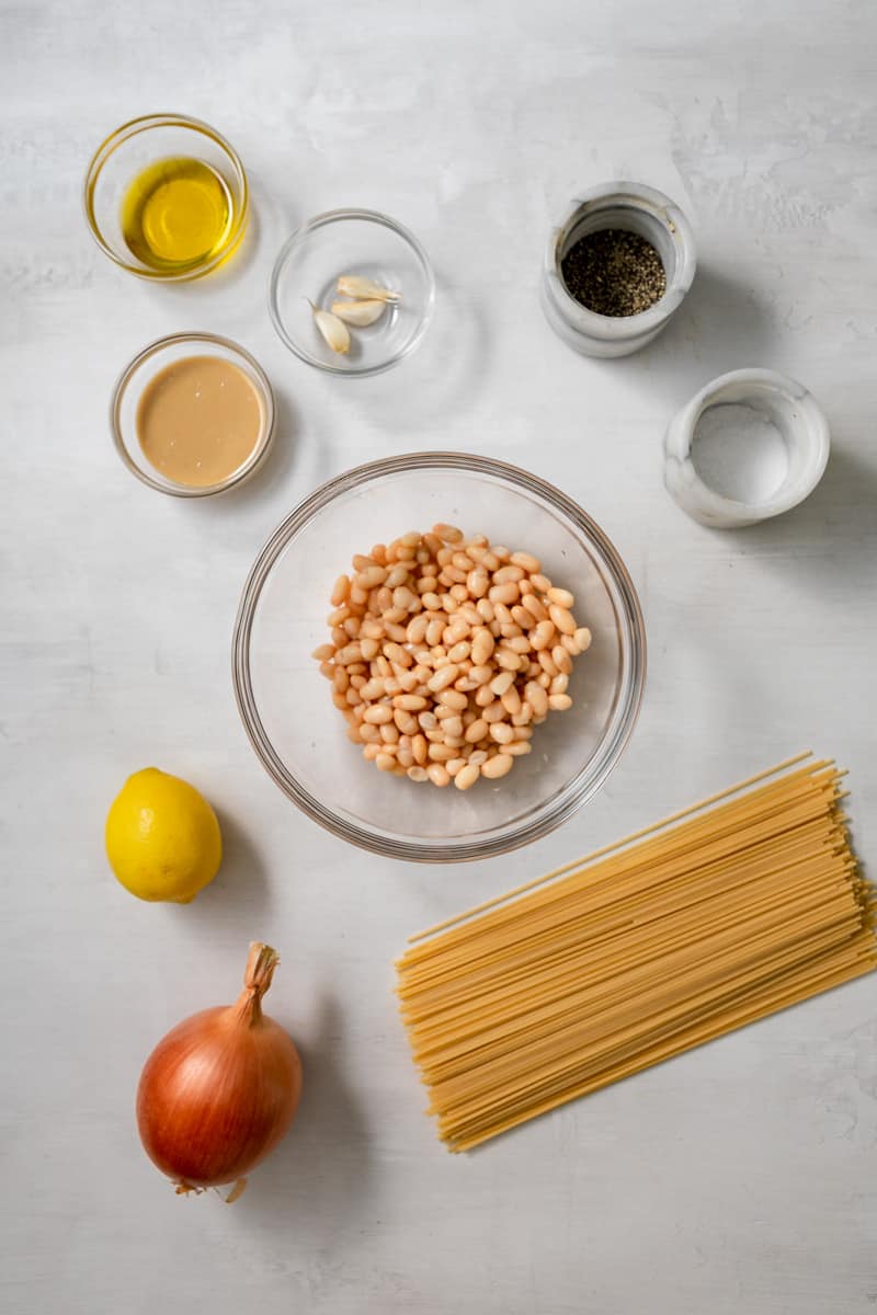ingredients in glass bowls: olive oil, tahini, garlic cloves, salt, pepper, white beans, lemon, yellow onion, dry spaghetti