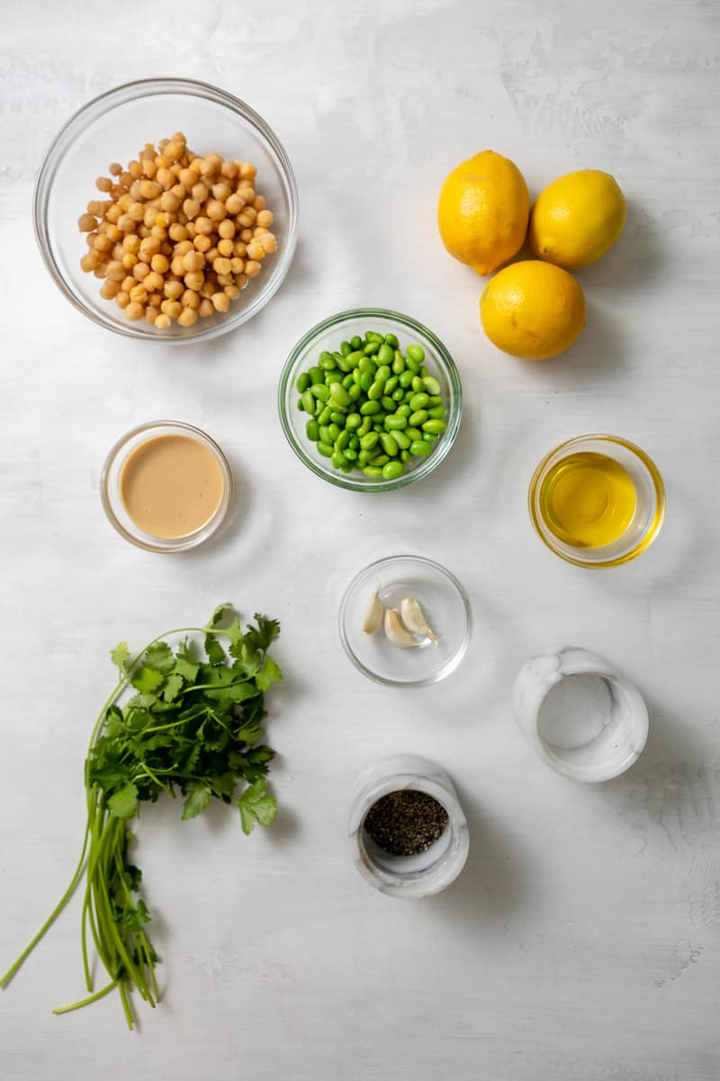 ingredients in glass bowls: chickpeas, lemons, tahini, edamame beans, olive oil, garlic, cilantro, salt, and pepper