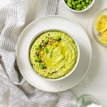 edamame hummus in a bowl on a plat topped with pine nuts, edamame beans and black sesame seeds