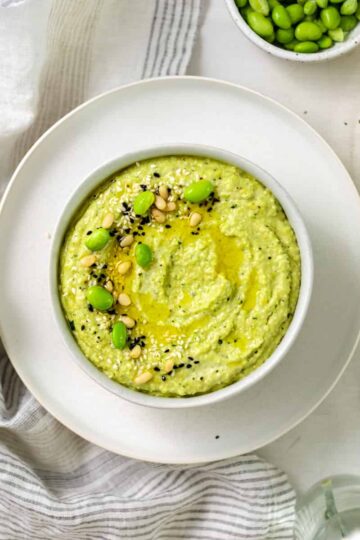 edamame hummus in a bowl on a plat topped with pine nuts, edamame beans and black sesame seeds.