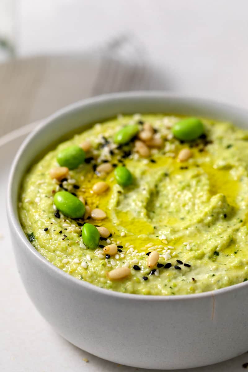 Bowl of edamame hummus up close, topped with pine nuts, edamame beans, black and white sesame seeds and a drizzle of olive oil.