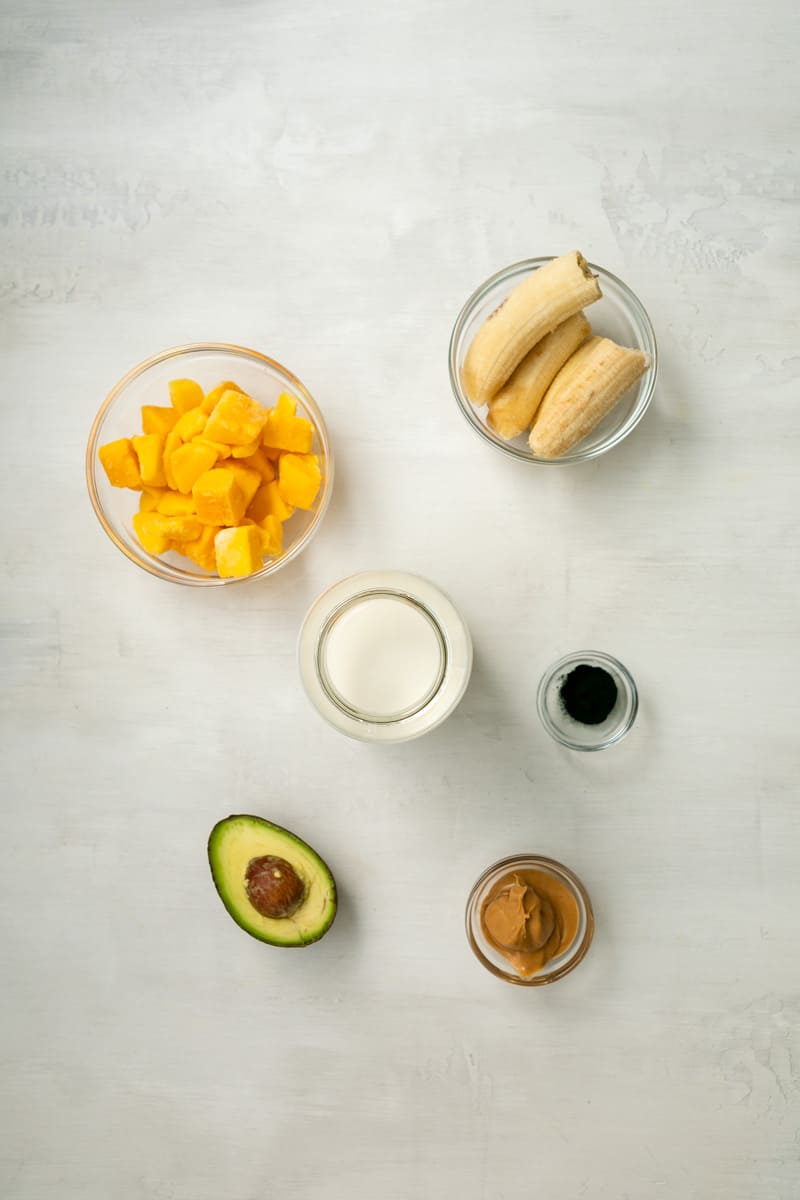 ingredients in glass bowls: frozen mango chunks, frozen banana halves, coconut milk, spirulina powder, half an avocado, peanut butter