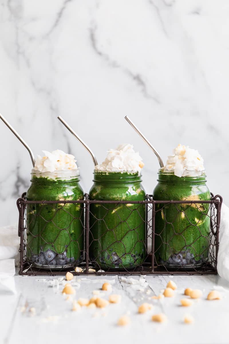 three mason jars with blueberries on the bottom, green smoothie, banana slices, then macadamia nut butter. topped with whipped cream, coconut flakes and more nuts, with a metal straw.