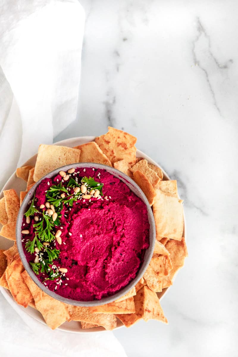 overhead shot of pink beet hummus in a bowl garnished with parsley and pine nuts, surrounded by pita chips