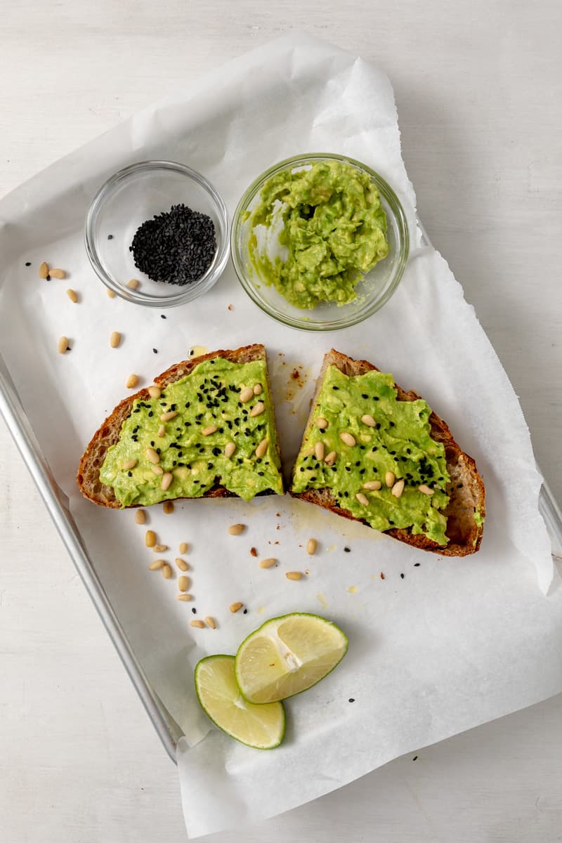 slice of avocado toast on a silver baking sheet lined with parchment paper, sliced in half topped with pine nuts and black sesame seeds. 