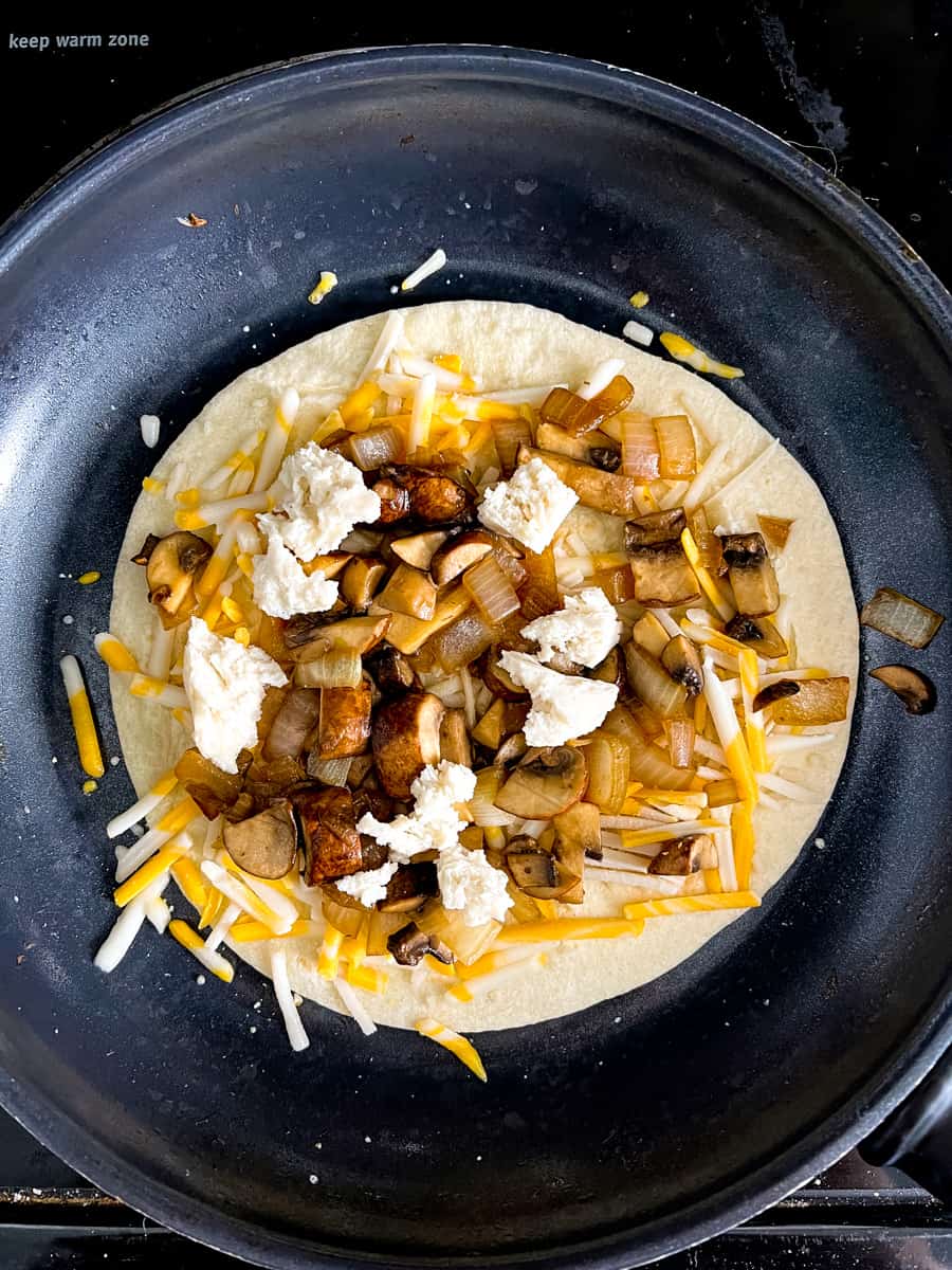 tortilla in a frying pan with mozzarella shreds, mozzarella chunks, sauteed onions and mushrooms and vegan cheddar jack shreds