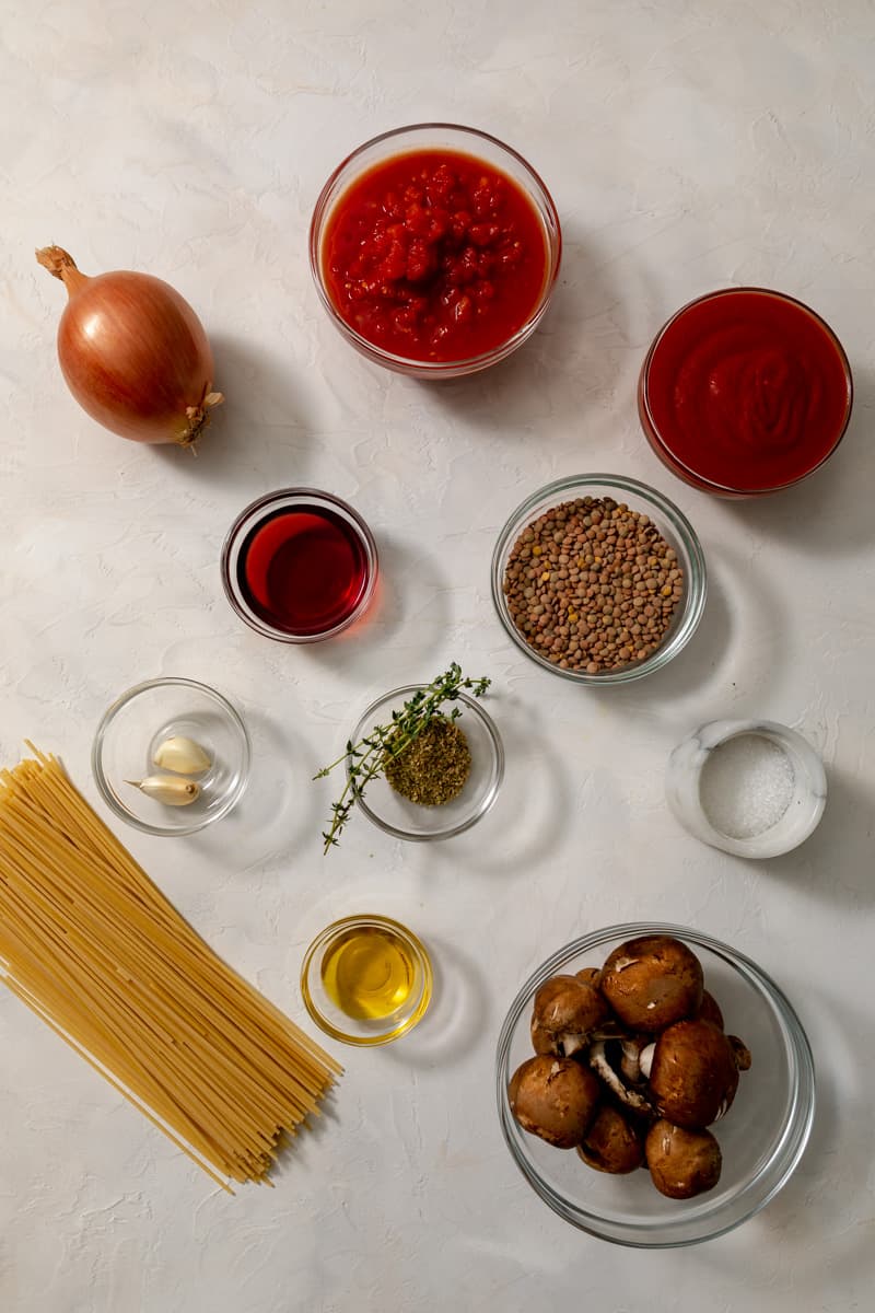 lentil spaghetti bolognese ingredients in glass bowls - onion, tomatoes, tomato sauce, red wine, lentils, garlic, thyme, oregano, salt, pasta, olive oil, and mushrooms.