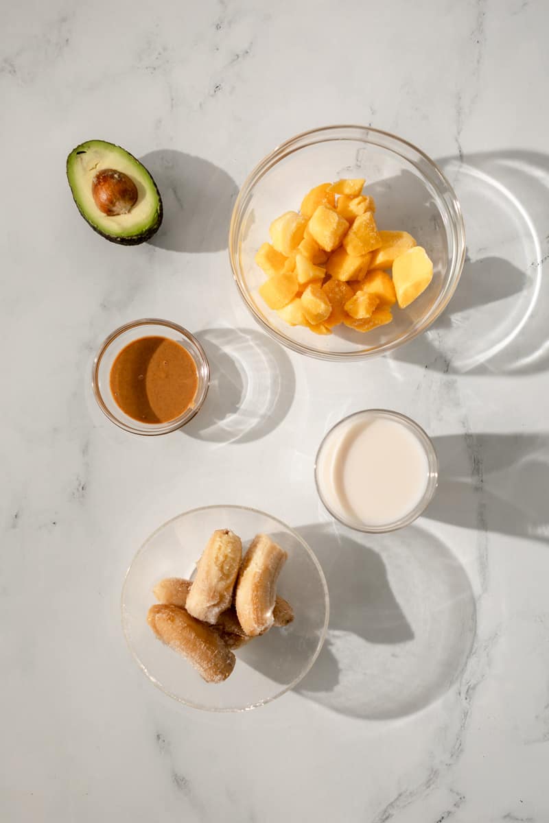 ingredients in glass bowls - avocado, frozen mango chunks, peanut butter, coconut milk and frozen banana halves.