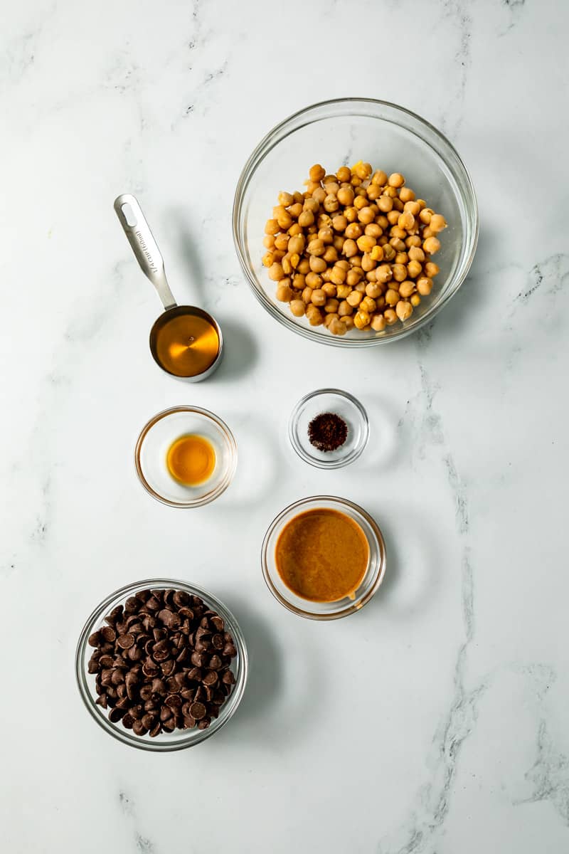 ingredients in glass bowls: chickpeas, agave, vanilla, coffee granules, peanut butter, and dark chocolate chips.
