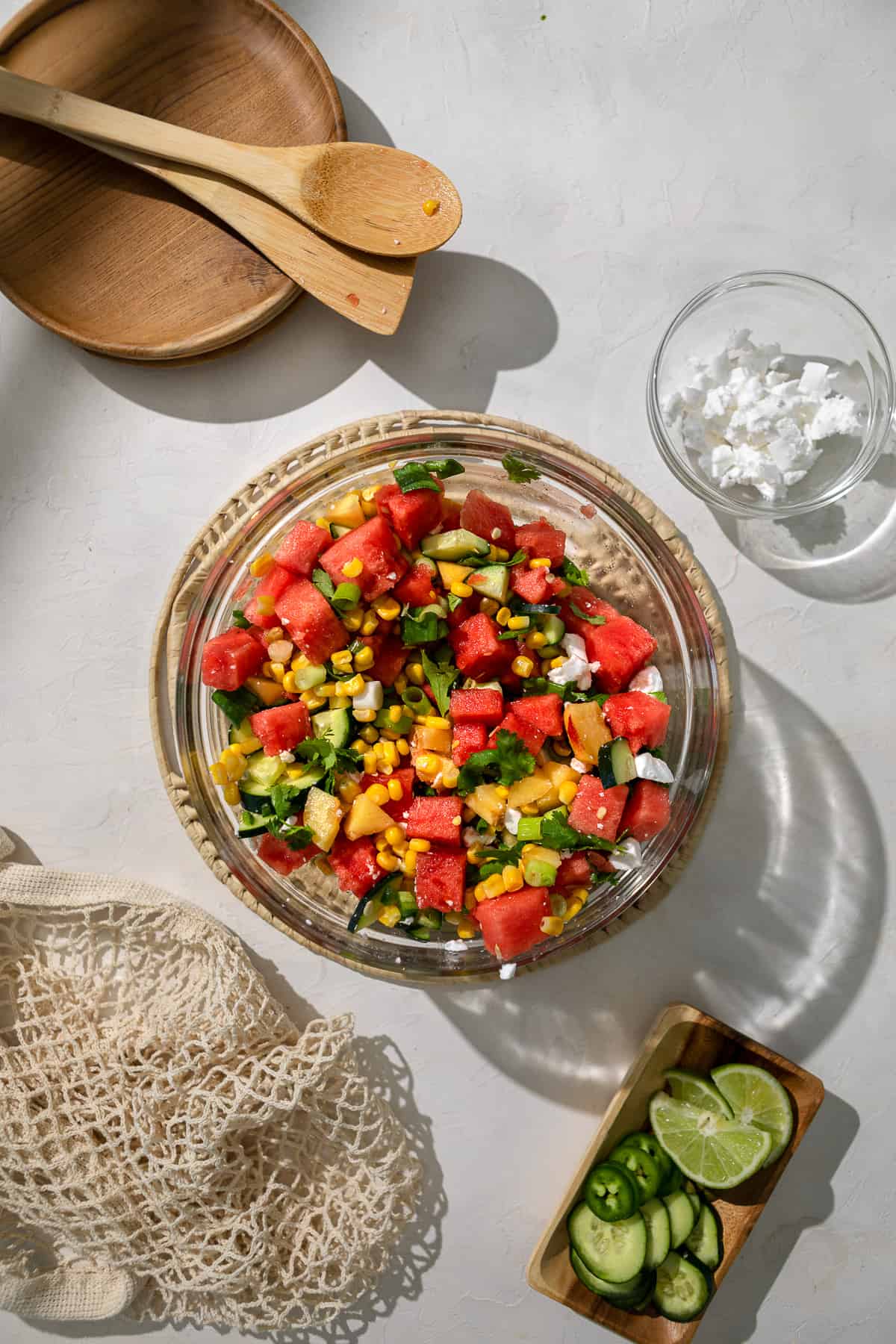 a glass bowl with watermelon, peach and corn salad, with wooden serving utensil and a wooden plate.